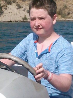 picture | Pulmonary Hypertension News | Cullen in a blue short-sleeve shirt, steering a boat. His collar is open, revealing a hint of a scar.