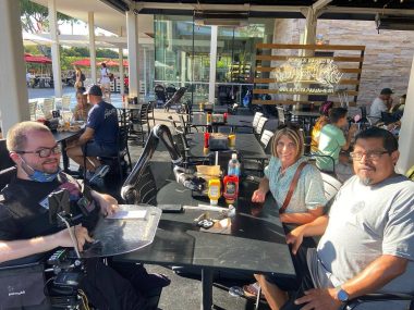 | Pulmonary Hypertension News | photo of a long dining table in a restaurant, with Jen and Manny on the right and Kevin, in a wheelchair, on the left