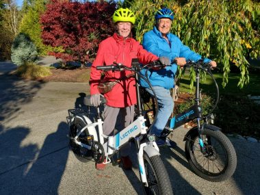 Carol and Dick Volckmann smile and pose with their e-bikes in the driveway, with a number of large trees behind them. Carol is wearing a red jacket, gloves, and a yellow helmet, while Dick is wearing a blue jacket and helmet. 