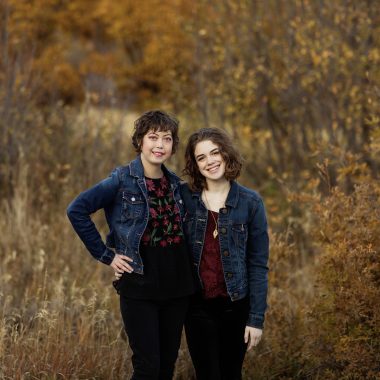 Two women, both wearing jean jackets, are standing in a field of brown. Both have dark hair and wear black pants. Both women have maroon in their shirts, though the shirt on the left also has black and the one on the right is totally maroon. The woman on the right wears a necklace.