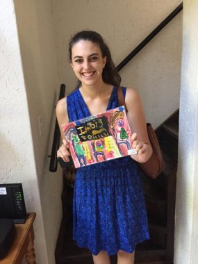 A young woman in a blue summer dress holds a piece of colorful children's art that says "I am big." She's standing in a stairway, smiling broadly.