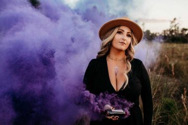 A woman with blond hair, a brown wide-brimmed hat, and a black dress, stands in a field filled with purple colored smoke. Her head is tilted slightly to the left, and she wears a contemplative look on her face. 