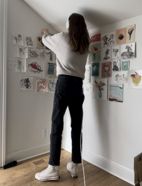 A woman stands, back to the camera, posting small artworks on paper to the wall. She has long dark hair and wears a gray shirt, black pants, and sneakers.