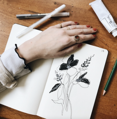 A sketchbook sitting on a brown surface is open to a page with a black-and-white ink drawing of flowers. A woman's hand is across the preceding page of the sketchbook, and a pencil, pens, and a tube are nearby.