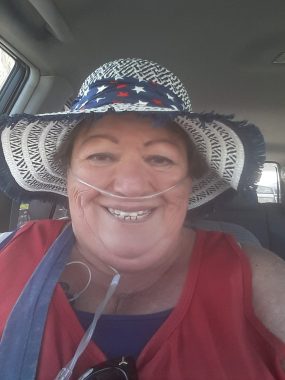 A head and shoulders photo of a woman in a car. She is using breathing tubes and wears a white and blue hat with a wide brim and a dark blue band with red and white stars. She has on a blue undershirt with a maroon sleeveless shirt over it.