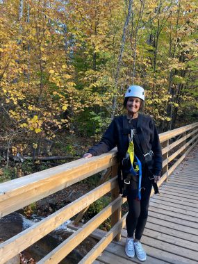 A woman poses on a bridge wearing a helmet and harness for zip lining. A river and fall foliage are visible in the background.