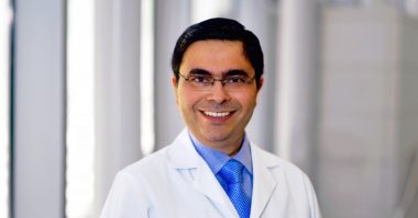 A professional headshot of a doctor. He has dark hair, glasses, and is wearing a white coat over a blue shirt and tie.