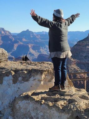 A woman stands on a rock with her arms spread, overlooking the Grand Canyon. She's wearing a beanie, a jacket, jeans, and boots.