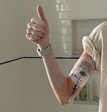 A close-up photo shows a woman's arm bent at the elbow and giving a thumbs up. A PICC line is coming out of her upper arm, covered with medical tape and bandages. The woman is wearing a white T-shirt with the sleeves rolled up and a watch. She's standing in front of a white tiled shower.