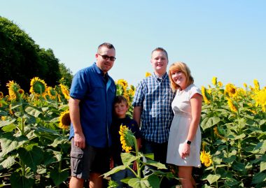 A family of four - two adults and two boys - stands in a sunflower field.