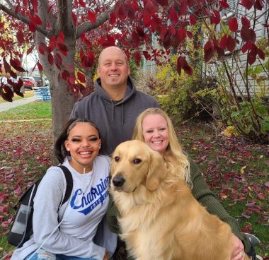 A family poses underneath the bright red leaves of a tree in autumn, in the yard of a home. A young woman, the daughter, is wearing supplemental oxygen; clockwise from her is a middle-aged bald man, a smiling woman, and a beautiful golden retriever that is starting straight into the camera and smiling.