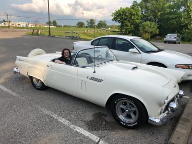 A woman sits in the passenger seat of an old, classic, cream-colored convertible. She's wearing glasses and a black shirt and has one arm resting on the door. The car is parked in a parking lot next to a more modern white car, and there are trees and a field in the background.