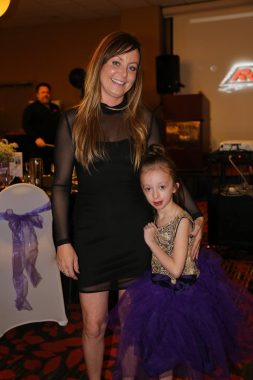 A woman in a short black dress with long dark blonde hair stands beside a small girl.