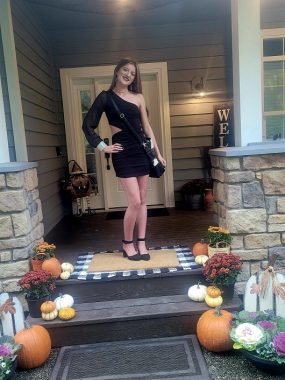 A young woman stands in a black cocktail dress on the front steps of a gray house with white. Halloween and autumn gourds are on the steps, including big and small pumpkins. 