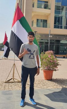 A young man stands outside in front of a United Arab Emirates flag. He has dark hair and is wearing a gray graphic T-shirt, dark pants, and blue sneakers. He is standing on a walkway in front of a tall apartment building with a large potted plant with pink flowers in the background.