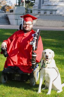 A young man with a beard and glasses and wearing a red university graduation gown and cap sits in a power wheelchair with a JACO robotic arm on the green lawn of a university. Next to him sits a large, white service dog with its mouth slightly open. The sun is shining brightly, casting shadows over the lower half of the man and on the lawn.