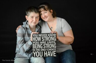 A portrait photo set to a black background shows a young mother who is white sitting next to and embracing her young son, who is also white. On their laps, they both hold a sign that says, "You never know how strong you are until being strong is the only choice you have."