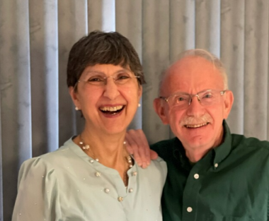 A photo shows a woman and man from chest up, standing before what appear to be white Venetian blinds. Both wear glasses. She has short-cropped dark hair with bangs and wears a pale green blouse and a necklace. He has gray, thinning hair and a mustached and wears a green button-down shirt.
