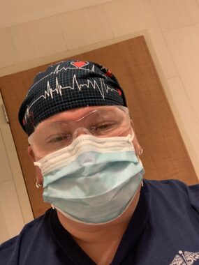 A nurse takes a selfie while working in the emergency department. She's wearing blue scrubs, a face mask, clear protective glasses, and a black cap with heart rate imagery.