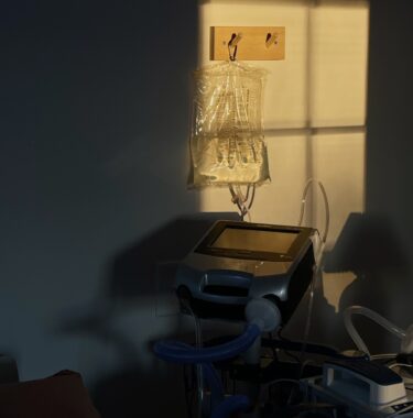 An image of Anna's bedside, featuring medical equipment. Most of the image is in shadows, but a square of sunlight highlights a humidification bag hanging on a hook just above a ventilator.