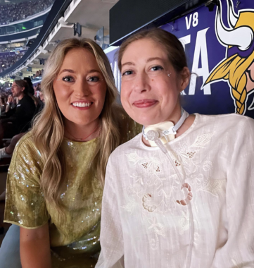 Two women who are sisters pose next to each other in a handicap-accessible section of a concert arena. A Minnesota Vikings logo is seen in the background.