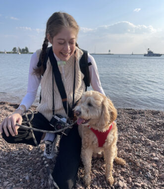On a pebble- or shell-covered shore with a large body of water behind her, a young woman (wearing long black pants and a white sweater vest over a long-sleeve white shirt) kneels next to a blond, midsize dog wearing a red harness. A breathing device and tube are just visible below the woman's chin, and a black strap around her neck connects to a camera about waist-high.