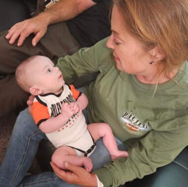 A photo taken from a high angle shows a woman holding her infant grandson. Her dark blond hair is tied back, and she's wearing a long-sleeve green T-shirt, while the baby is wearing a Harley Davidson onesie. Both grandma and grandson are looking at each other and smiling.