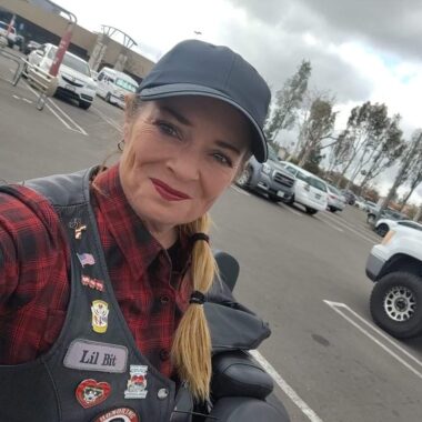 A woman takes a selfie next to her motorcycle in what appears to be a shopping center parking lot. She's wearing a black cap, buffalo plaid shirt, and a black leather vest with various patches and pins.