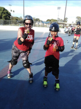 A woman and her son wear matching red T-shirts at a roller skating event. Both have on helmets, shoulder and knee pads, and roller skates.