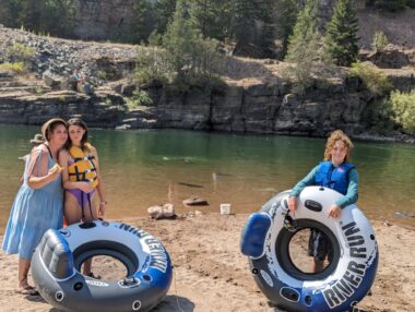 A mother, her young daughter, and a teenage boy stand next to a river with bluffs in the background. The kids are holding large inner tubes for floating down the river. The daughter and the boy are wearing life jackets. It's a sunny day. 