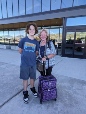 A mother and son stand in front of an airport. The son looks like a teenager, dressed in blue shorts and a blue T-shirt. He has wavy, chin-length hair, and stands about a foot taller than his mother, who's at his left. She holds the handle of a purple and black suitcase.