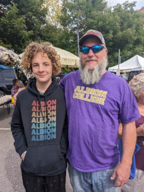 A bearded man wearing a baseball cap and sunglasses stands next to his son, a teenager with curly hair. The man is wearing a purple T-shirt and the son a black hoodie. They seem to be at an outdoor fair. 