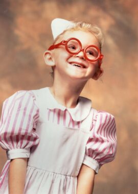 A professional photo shows a young girl against a light brown backdrop. The girl is dressed as a nurse, in a red and white striped dress, white smock, white cap, and red glasses.
