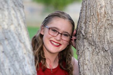 A professional, close-up photo shows a high school girl smiling and posing between tree trunks. She's wearing a red shirt and glasses and is smiling at the camera.