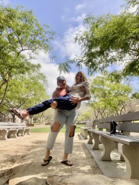 A low-angle photo shows a young adult woman holding an older woman in her arms. They appear to be in a park, between rows of benches and surrounded by trees. Both women are looking at the camera and smiling broadly.