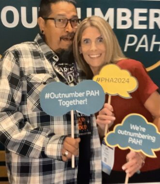 A man with a black-and-white checkered shirt, a black T-shirt, and glasses stands beside a woman with long blond hair and a red shirt. They hold signs with quote balloons that read "#Outnumber PAH Together!," "#PHA2024," and "We're outnumbering PAH!" They're in front of a dark green wall or poster that seems to include part of the words "OUTNUMBERING PAH."