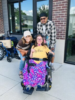 Three adults pose for a photo on the sidewalk in front of a coffee shop, whose exterior features bricks and large windows. The woman in the center is wearing a yellow T-shirt and seated in a power wheelchair. Another woman and her husband stand just behind her and are leaning in and smiling.