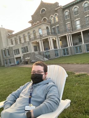 A young man in his 20s sits on a white Adirondack chair on the lawn in front of a college building. He's wearing a gray sweatshirt, a black face mask, glasses, and has a lanyard around his neck.