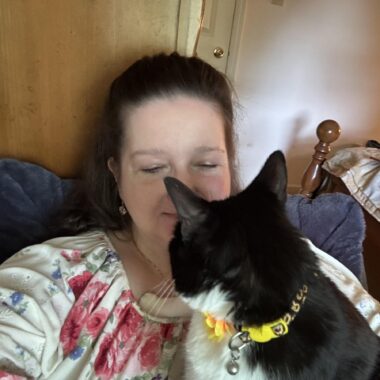 A woman has a black and white cat on her lap as she sits in what appears to be a blue chair in a home. She wears a white blouse with blue and red flowers; the cat has a yellow collar.