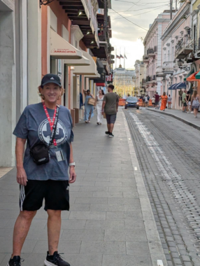 A woman pauses for a photo on a sidewalk in a town on one of the Caribbean islands. She's wearing a T-shirt, athletic shorts, baseball cap, and sneakers, and has a lanyard around her neck and a small cross-body bag looped over one shoulder.