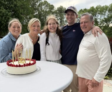 A family of five - two daughters, one son, a father, and a mother - stand around a circular outdoor table with a birthday cake on it. The five have their arms around one another.