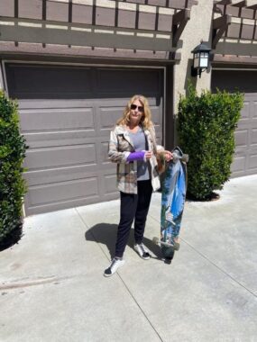 A woman stands in a driveway on a sunny day. She has a purple cast on her right wrist and holds the top of a long skateboard in her left hand, while the bottom of the skateboard rests on the ground. She's wearing sunglasses.