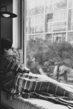 A woman wrapped in a striped blanket sits on a window seat, gazing outside at a rainy scene with buildings and trees visible through the wet glass. The image is in black and white.