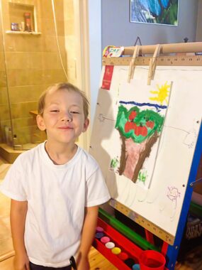 A young boy in a white T-shirt smiles proudly next to an easel displaying his colorful painting of a tree with red apples, green leaves, a blue sky, and a yellow sun. Art supplies are visible on the easel’s tray.