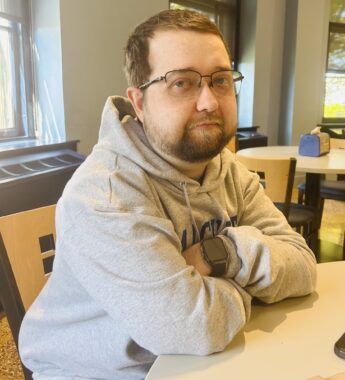 A 25-year-old man sits at a round table with his arms crossed in front of him. He's wearing a gray sweatshirt, glasses, and a smartwatch, and is looking to his right toward the photographer.