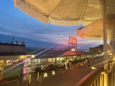 View of Pike Place Market at dusk, with a glowing neon sign, cobblestone street, and market stalls. Large white umbrellas with fringe frame the scene from above, and the sky is tinged with blue and pink hues.