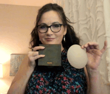 A woman with shoulder-length brown hair and brown or black glasses holds a square black box in one hand and a beige bra cushion in the other. 