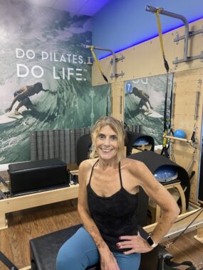 A woman in a black tank top and blue leggings sits on a reformer machine in a Pilates studio. She has one hand on her hip and is smiling happily at the camera.