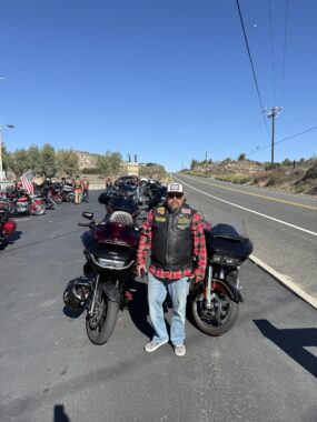 A man in a red plaid shirt, black leather vest, jeans, and a hat stands in front of several motorcycles parked next to a road.