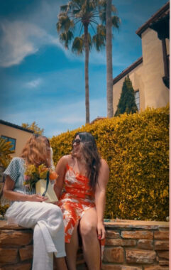 Two women, a mother and daughter, sit on a stone bench and smile happily at each other. The mom is holding a bouquet of flowers, and a palm tree towers over them in the background.